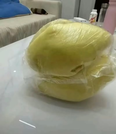 图中白色光滑桌面上放置着用透明塑料袋封装的两块淡黄色馒头，其外形、色泽和剥好的榴莲果肉高度相似，很容易让人产生误认，背景可见浅色的布艺沙发以及摆放在桌面边缘的水瓶、杯子等家居物品，整体是居家场景下拍摄的趣味实拍图。