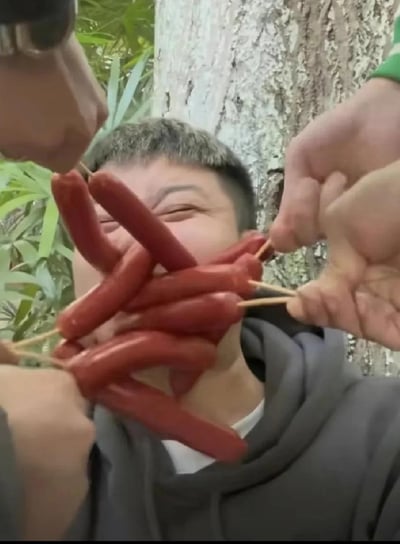 这是一张户外整活的照片，一名短发男子被多根串在竹签上的小香肠凑到脸前，眼睛挤成一团，露出哭笑不得的表情，周围的人拿着串着香肠的竹签在他脸前摆弄，是朋友间的恶作剧互动，背景是树木和绿植，氛围轻松欢乐，没有双关或谐音梗内容。