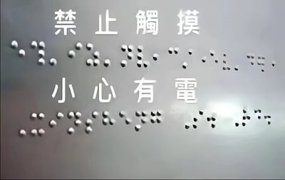 图片展示了一个警示标识，上方有白色中文文字'禁止觸摸'，下方有'小心有電'。文字下方对应排列着凸起的盲文点字，整体背景为灰色金属质感，光线从右侧照射形成轻微反光效果。这是一个标准的电气安全警示标志，用于提醒人们不要触摸相关设备以防触电。