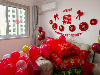 这是一间布置完成的中式婚房，墙面装饰着红色的“新婚大喜”字样、巨型“囍”字，搭配红色纸扇、立体蝴蝶结、金色蝴蝶贴纸，还有一条写着“HAPPY WEDDING”的红色拉花；床上铺着带有龙凤呈祥、百子闹春传统纹样的红色婚被，床的周围摆放着红色、金色、透明带金箔的气球，部分气球印有“囍”字；窗户玻璃上贴着红色的喜字窗花，窗外是高层居民楼，整个空间充满了中式婚礼独有的喜庆与温馨氛围，处处彰显着新婚的喜悦感。