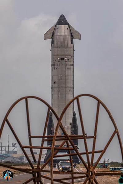 画面中是SpaceX研发的星舰（Starship）测试原型舰，正被大型转运设备承载着在道路上移动，前景的金属框架形成类似心形的视觉遮挡，背景是美国得克萨斯州博卡奇卡Starbase测试基地的相关设施，天空阴沉，整体场景展现了重型星际运载工具研发过程中的硬核一幕。