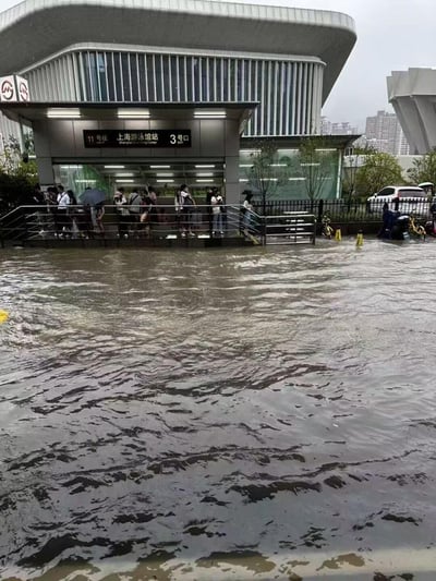 这张图拍摄的是上海地铁11号线上海游泳馆站3号口，因强降雨导致周边路面严重积水，浑浊的积水几乎漫到站口台阶处，大量乘客聚集在站口无法出行，部分乘客打着伞，现场能看到积水在路面流动，还有被淹的非机动车，呈现出城市内涝后的窘迫场景。