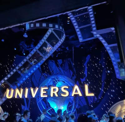 夜晚的环球影城标志性入口，巨大的黄色"UNIVERSAL"字样在蓝色灯光下格外醒目，背景是一个发光的蓝色地球仪，两侧装饰有电影胶片造型的结构，胶片上能看到模糊的电影画面。画面下方有许多游客聚集，整体氛围梦幻而充满电影感。