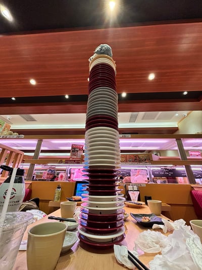在一家日式寿司店内，餐桌中央堆叠着一座惊人的餐盘塔，红白相间的寿司碟层层叠起，几乎触及天花板。塔顶还放置了一个小型毛绒玩具作为装饰。周围可见典型的寿司店环境，包括木质装饰、柜台座位和菜单看板，桌上散落着饮料瓶、茶杯和用餐后的残留物，展现出一场丰盛的寿司大餐后的场景。