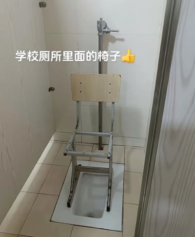 这张图片展示了学校厕所隔间内的场景。画面中可见白色瓷砖铺就的墙壁和地面，中央是一个白色的蹲便器，上方被一把木质座椅面、金属支架的椅子覆盖，椅子四条腿分别位于蹲便器四周。隔间右侧有一个银色的冲水阀。图片上方有白色文字标注“学校厕所里面的椅子👍”，并配有一个点赞表情，推测是为了方便使用蹲便器而进行的简易改造，带有一定的实用性和校园生活气息。