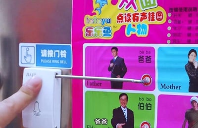 这是一张创意拼贴的搞笑图片，画面左侧是一个正在被手指按下的白色门铃，门铃的金属推杆恰好对准右侧的乐乐鱼双面点读有声挂图中“爸爸”板块的触发按钮，形成了“按门铃就会发出‘爸爸’的声音”的错位巧合，制造出无厘头的幽默效果。右侧的点读挂图是儿童认知亲属的教具，上面印有“爸爸”“妈妈”“伯伯”等亲属的真人图片、中文称呼、拼音及英文翻译，还有按键使用说明，整体场景将日常门铃和儿童教具进行趣味结合，充满反差感。