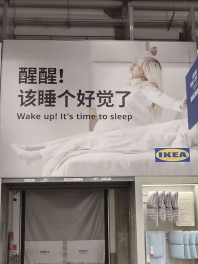 这是一张拍摄于宜家线下门店的实拍照片，画面上层是宜家投放的睡眠相关产品宣传海报：海报右侧是一名金发女性模特坐在白色床品上、刚睡醒伸懒腰的形象，左侧搭配反差感文案“醒醒！该睡个好觉了”，下方对应英文翻译“Wake up! It's time to sleep”，海报右下角标注有宜家的蓝黄配色品牌LOGO。海报下方可见门店的出入口区域以及侧面陈列的家居布艺产品，文案用看似矛盾的表达，调侃当代人作息不规律、被琐事消耗需要停下来好好休息的现状，极具网感和趣味性。