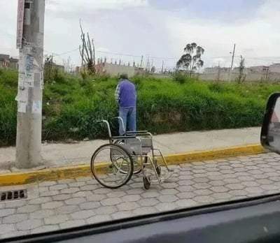 照片中，一名身穿紫色夹克和蓝色牛仔裤的男子站在人行道旁，身旁停放着一辆银色轮椅。男子背对着镜头，低头望向路边的草地。画面从车内视角拍摄，可见车窗边框和部分侧后视镜，背景是长满青草的斜坡、贴有海报的电线杆、几棵树木及远处的低矮建筑群，天空多云。轮椅的存在与男子站立的状态形成视觉反差，带来“本以为需要轮椅的人却能站立”的意外感。