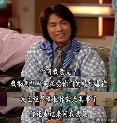 图片为香港电视剧《男亲女爱》的剧照截图，演员黄子华饰演的陈十三穿着蓝白格子睡衣，内搭衬衫领带，坐在沙发上，脸上带着略显僵硬的微笑。画面中叠加了四行文字：“你问我意见？我很明显就是在受你们的精神虐待 我已经尽量装作若无其事了 你还走过来问我意见”。通过角色强装镇定的表情与内心委屈的文字形成反差，营造出无奈又搞笑的效果，常被用于表达明明内心不满却不得不表面平静的场景。