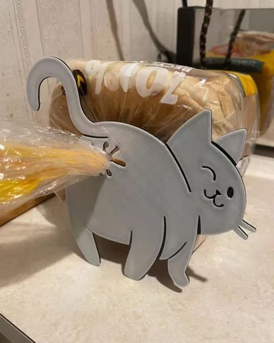 厨房台面上摆放着一个灰色卡通猫咪造型的面包架，猫咪面带微笑，尾巴向上翘起。面包架中间放置了一袋切片面包，面包片从猫咪背部的开口处‘挤’出来，形成猫咪‘排泄’面包的趣味视觉效果。背景中可见部分包装食品，整体场景展现了这款创意家居用品的可爱与幽默设计。