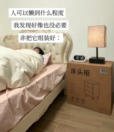 这张实拍图展示了居家卧室场景，左侧是带有雕花的欧式软包床，床上有人盖着被子躺着休息，床的右侧本应放置床头柜的位置，直接摆放了还没拆封、未组装的床头柜原包装箱，包装箱的顶面被直接当作台面用来放置台灯和显示时间为8:18的电子时钟。图上方配有三行文字，用自嘲的口吻调侃极致偷懒的行为：不需要费心组装床头柜，直接用外包装箱就能实现床头柜的摆放功能，笑点十足。