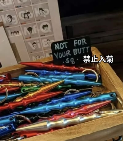 图片展示了一个商店的趣味商品陈列：一个木质盒子里装满了彩色金属钥匙扣，这些钥匙扣形状类似小匕首或尖刺。盒子上立着一个黑色小标语牌，用白色字体写着"NOT FOR YOUR BUTT $8"，旁边配有中文翻译"禁止入菊"。商品定价8美元，标语通过幽默方式暗示了这些尖刺状钥匙扣的正确使用方式，避免消费者产生不当联想。背景中可以看到墙上挂着一些人物头像的装饰画。