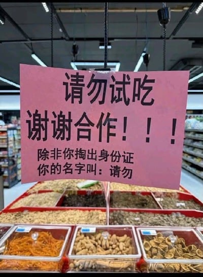 超市货架区悬挂着一块粉色标语牌，牌面上方用黑色大字写着“请勿试吃 谢谢合作！！！”，下方却以幽默口吻补充“除非你掏出身份证 你的名字叫：请勿”。通过将“请勿”二字曲解为姓名，形成文字游戏，制造出反差萌的搞笑效果。背景可见超市陈列的各类坚果、干货等散装食品，灯光明亮，整体场景为真实的超市购物环境。