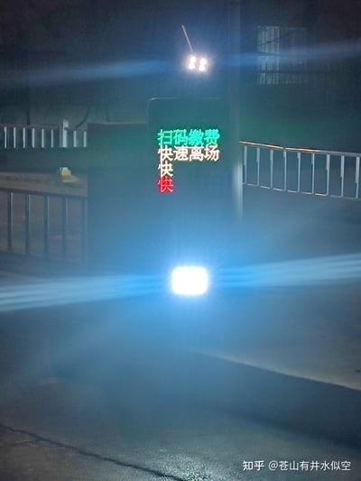 这是一张夜晚拍摄的停车场出口实拍图，画面中央的立柱上设有电子发光提示牌，依次显示绿色的「扫码缴费」、白色的「快速离场」以及红色的「快快快」，下方有车辆的强光车灯在画面中形成亮斑，背景可见停车场的金属栏杆与建筑墙体，整体环境昏暗，提示牌的彩色发光文字在夜色中十分醒目，用于催促车主完成缴费后快速离场。