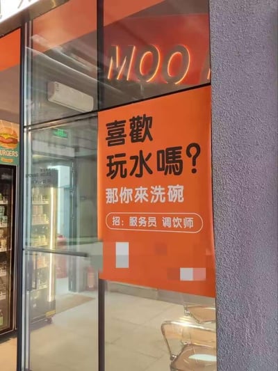 图片展示了一家店铺的玻璃外墙，上方有橙色发光的“MOO”字样招牌。墙面中央张贴着橙色背景的招聘海报，海报以黑色大字醒目地写着“喜歡玩水嗎？”，下方紧接着用白色小字回应“那你來洗碗”，底部则正式标注“招：服務員 調飲師”。海报通过幽默的语言将“玩水”与“洗碗”巧妙关联，以轻松诙谐的方式吸引求职者注意。背景中可隐约看到店铺内部的冰柜和桌椅，左侧玻璃上还能看到“BURGERS”的字样，推测可能是一家汉堡或饮品店。