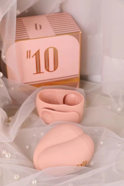 画面展示了一组粉色系产品，主体为一个印有金色"10"字样的粉色礼盒，礼盒设计带有横向条纹装饰和金色边框。礼盒前方摆放着两个粉色容器，一个是打开的椭圆形收纳盒，另一个是闭合的心形盒子。整个场景铺设着白色纱幔，点缀着珍珠装饰，营造出精致优雅的氛围，可能是美妆或奢侈品的产品展示图。