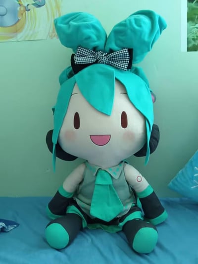 这是一张手工制作的超大号初音未来毛绒玩偶的实拍照片，玩偶还原了初音未来标志性的青绿色发型、黑色耳机、公式服造型，还额外添加了黑白格纹蝴蝶结装饰，玩偶面带开心的笑容，脸颊带有红晕，端坐于蓝色床面之上，背景是浅绿色墙面，整体风格软萌治愈，充满手工制作的温度。