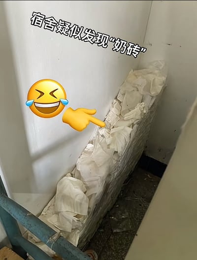 图片展示了宿舍墙角堆积的白色物体，看起来像整齐堆叠的'奶砖'，实际上是大量白色纸巾或纸张被塞进角落形成的有趣视觉效果。图片左上角配有文字'宿舍疑似发现"奶砖"'，旁边有一个笑到流泪的表情符号手指着这堆"奶砖"，营造出幽默搞笑的效果。