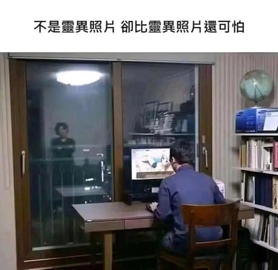 图片展示了一个办公室或书房场景，一名男子正专注地看着电脑屏幕。通过他身后的玻璃门，可以看到一个女人的身影站立着，营造出类似恐怖片中常见的"灵异照片"效果。图片上方配有文字"不是灵异照片 却比灵异照片还可怕"，暗示男子正在看的内容比身后的人影更加令人恐惧，形成一种黑色幽默的反差效果。