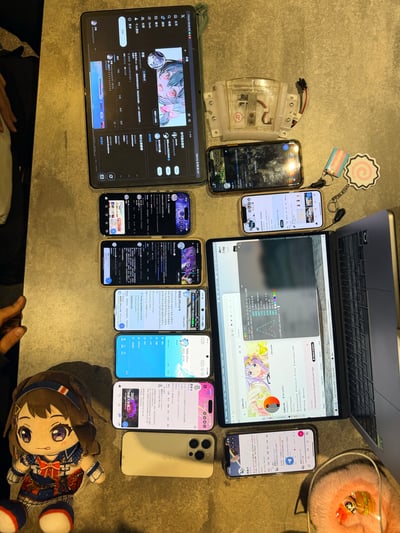 一张俯拍的桌面照片，展示了丰富的桌面场景：中央及右侧摆放着1台笔记本电脑、1台平板电脑和至少8部智能手机，设备屏幕亮着，显示聊天记录、动漫图片、代码界面等不同内容。左下角有一个穿着水手服的卡通女孩玩偶，有着紫色大眼睛和棕色短发，表情可爱。桌面上还散落着透明塑料零件、带挂饰的钥匙链、浅色手机壳及粉色毛绒物品，整体呈现出科技产品与二次元元素结合的生活化场景，给人一种充实且略带杂乱的日常感。