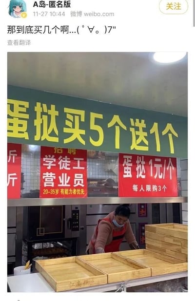 这是一张来自微博的社交媒体截图，顶部是微博用户“A岛-匿名版”发布的动态，配文“那到底买几个啊...(゜∀。)7"，直白地表达了对下方店铺促销规则的困惑。截图内嵌的实拍图展示了一家名为“江南糕点”的线下糕点店柜台区域：柜台上方悬挂着一块醒目的绿色标语，写着“蛋挞买5个送1个”；右侧的红色告示牌则标注“蛋挞1元/个 每人限购3个”，这两个蛋挞促销规则明显相互冲突，一个要求购买5个才能享受赠送，另一个却限制每人最多购买3个，形成了无法同时满足的矛盾要求，让消费者不知如何选择。柜台中间的红色告示牌是招聘信息，内容为“招聘 学徒工 营业员 20-35岁有能力者优先”；左侧红色告示牌仅露出部分“斤”字，内容不完整。柜台前，一位穿着粉色上衣、红色围裙并佩戴口罩的女性工作人员正在整理几个空的木质托盘，柜台后方可以看到烘焙烤箱等设备，整体场景是日常的糕点店经营环境。