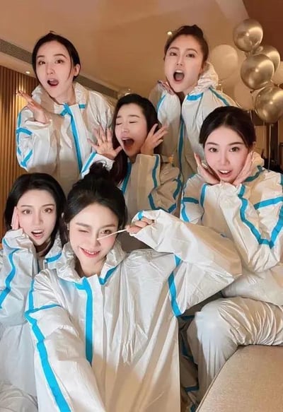 这是一张室内拍摄的静态合影照片，画面里的六位年轻女生身着带有蓝色装饰线条的白色防护服，在布置有银色、白色气球的室内环境中摆出各式各样的搞怪姿势与表情：有的张大嘴巴露出惊讶神态，有的双手做出可爱托脸动作，还有一位女生手持类似核酸采样棉签的道具，眯起一只眼睛做出俏皮的样子，整体氛围轻松欢快，应该是疫情背景下好友间的趣味留影，图中没有双关、谐音梗或其他暗示内容。
