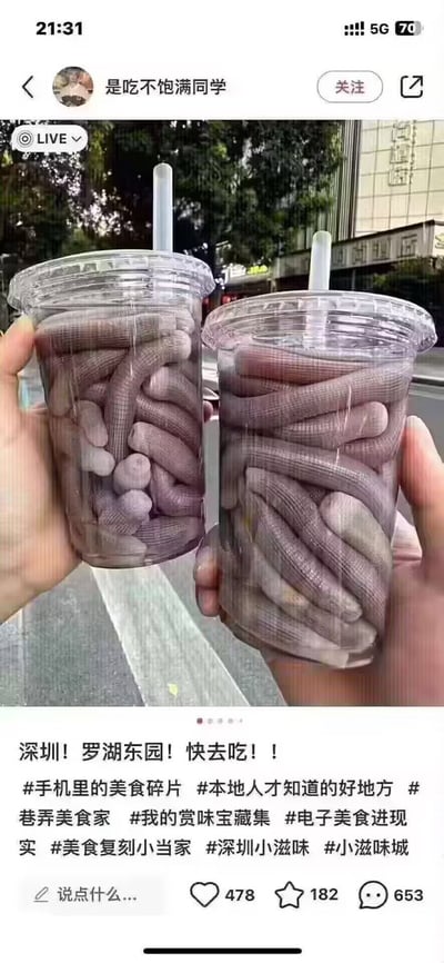 这是一张社交媒体内容截图，画面主体是两只手分别握持两个带盖、插有吸管的透明奶茶杯，杯中并没有常规奶茶饮品，而是塞满了灰紫色的沙虫（又称泥丁、海肠），视觉上仿佛是特殊的“沙虫奶茶”，背景是户外城市街道场景。截图下方是发布者配文“深圳！罗湖东园！快去吃！！”，附带多个美食相关的话题标签，页面显示该内容获得478点赞、182收藏、653条评论，发布者账号名为“是吃不饱满同学”。该内容用奶茶杯盛装沙虫的形式制造出强烈的视觉反差，引发网友讨论。