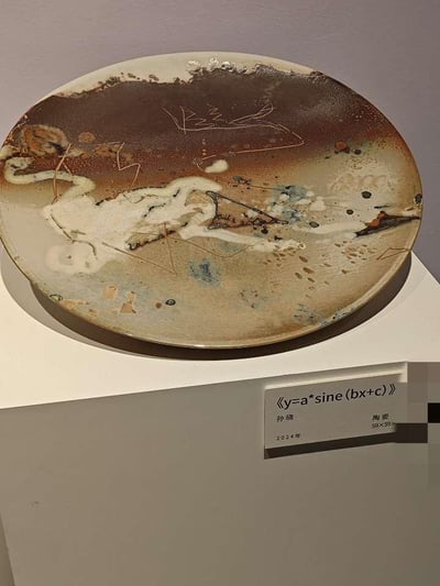 这是一张艺术展览现场的实拍照片，展示了艺术家孙晓2024年创作的陶艺作品《y=a*sine(bx+c)》。作品为圆形陶艺盘，盘面以抽象风格呈现：上部区域是深棕色调的窑变釉面，带有自然形成的肌理质感；中部及下部以浅米色釉色为底，点缀着白色、蓝灰色、深褐色的釉彩斑块与泼溅痕迹，同时用纤细的金色线条勾勒出类似函数曲线的形态，将数学函数的理性概念与陶艺窑变的感性视觉效果相结合，营造出写意又富有思辨感的艺术氛围。作品下方的白色说明牌清晰标注了作品名称、作者孙晓、创作年份2024年、材质陶瓷以及尺寸信息，整体场景简洁，突出了陶艺作品本身的艺术表现力。