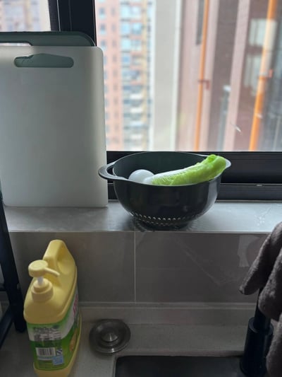 厨房窗台上的备菜场景：黑色滤水碗中放着一颗白菜和两个鸡蛋，旁边是白色塑料砧板，下方台面上有黄色洗洁精瓶、金属水槽塞和灰色毛巾，窗外可见模糊的城市高楼背景。整体呈现出日常家庭烹饪前的准备状态，充满生活气息。