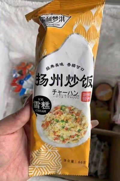 这张图片展示了一只手拿着瑞利梦淇品牌的扬州炒饭口味雪糕包装，包装主色调为暖黄色，中间区域印有一碗色泽丰富的扬州炒饭实物图，搭配“经典美味 香糯可口”的宣传语，同时标注了日文“チャーハン”，明确产品为“雪糕”，净含量66克，生产日期为2023年4月9日，还带有“新鲜营养”的标识。这款雪糕将中式经典主食扬州炒饭与冷饮雪糕结合，属于市场上的猎奇跨界创新食品，打破了大众对雪糕口味的常规认知，给人强烈的新奇感与反差感，无谐音梗、双关或暗示类内容，核心亮点在于口味的跨界创意。