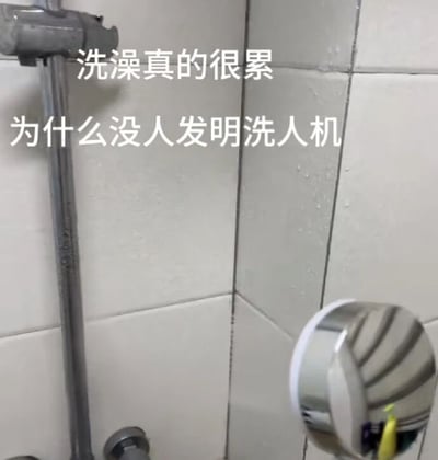 图片展示了一个浴室角落的实景，墙面铺着白色瓷砖，左侧有金属水管和淋浴装置，右下角可见银色淋浴喷头。图片中央叠加白色文字：“洗澡真的很累 为什么没人发明洗人机”，表达了对洗澡过程感到疲惫的日常吐槽，并幽默地提出希望有类似洗衣机的“洗人机”发明来解决这一“烦恼”。内容贴近生活，充满轻松诙谐的气息。