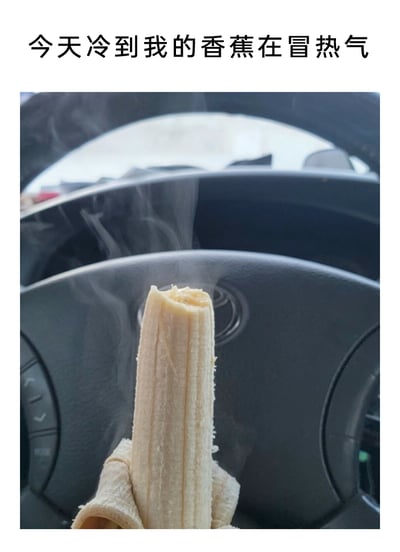 这是一张用来调侃极寒天气的搞笑创意图，画面背景是汽车的方向盘，前景是一根剥了皮的香蕉，香蕉周围环绕着类似热气的白雾。搭配上方的文字“今天冷到我的香蕉在冒热气”，用夸张的手法将天气寒冷的程度具象化，仿佛常温的香蕉在低温环境下都因“温度差”而冒出热气，通过日常食物的趣味场景制造诙谐效果，表达出对严寒天气的调侃。