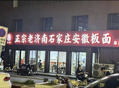这是一张夜晚拍摄的街边实拍静态图，核心内容是一家板面店的红色发光招牌，招牌上标注“正宗老济南石家庄安徽板面总店”，将分属三个不同省份的地名组合在一起，违背了安徽板面是安徽特色美食的常规认知，充满违和感和笑点。画面中店铺为临街底商，玻璃门窗内可以看到有多名顾客正在用餐，店外人行道上停放着电动车、共享单车，还有行驶的电动车和停放的私家车，画面左侧还有禁止停车的交通标识，整体是充满烟火气的城市街边日常场景。