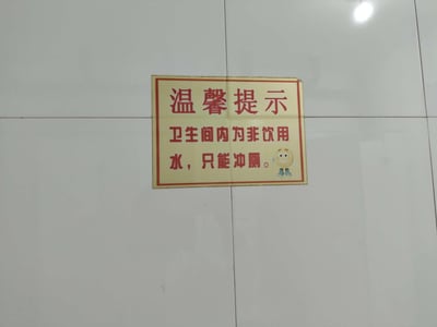 一张贴在白色瓷砖墙上的温馨提示牌，牌面为黄色底红色边框，内容用红色字体写着“温馨提示 卫生间内为非饮用水，只能冲厕。”，末尾配有一个戴着蓝色鞋子的笑脸表情符号。提示牌旨在明确告知卫生间用水的用途限制，语言简洁直接，搭配表情符号增添一丝轻松感。