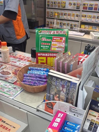 便利店收银台场景，货架上摆放着各种商品，显眼处有2024圣诞购物节促销海报，写着"终极价值2500点"、"天天送100元"等促销信息。柜台上有一篮Mentos糖果，旁边放置着一本封面文字极具争议的书籍，内容为"就以为有钱的客人了不起 找个地方默默地去死"，形成普通商业场景与极端言论的强烈反差。