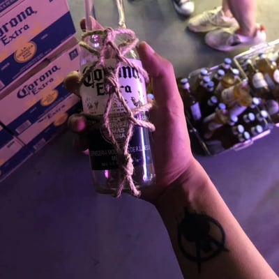 一只手握着一瓶用麻绳缠绕的科罗娜特级啤酒，瓶身上可见品牌标识和"La Cerveza Mas Fina"字样。背景中堆叠着多个科罗娜啤酒箱，右侧有装满啤酒瓶的透明塑料箱，地面上能看到几双穿着休闲鞋的脚，暗示这是在聚会或社交场合拍摄的场景。手臂上有一个黑色纹身图案。