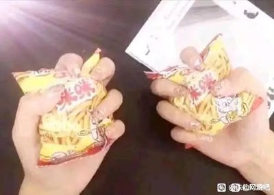 这张静态单图展示了深棕色桌面上，一左一右摆放着两只人手，每只手各握着一包被捏皱的黄色包装咪咪虾条，画面右上角露出一张带有黑色图案的白色纸张，图片右下角带有百度贴吧的水印。该图利用咪咪虾条名称里的“咪咪”一词构成谐音玩梗。