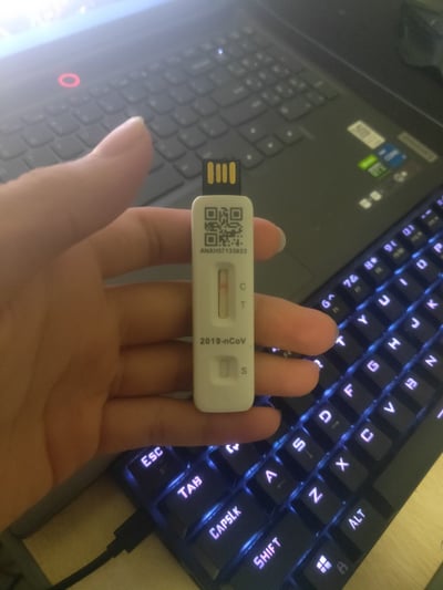 这是一张实拍照片，画面中一只手捏着一个外形完全模仿新冠抗原检测试剂的U盘进行展示，U盘顶部是标准USB公头接口，主体部分印有新冠抗原检测试剂常见的二维码、产品编号、C/T/S检测标识以及“2019-nCoV”字样，仅从外形看极易被误认为是真正的抗原检测试剂。背景可以看到带背光的机械键盘和一台笔记本电脑，整体是在桌面数码使用场景下拍摄，凸显这款U盘的恶搞创意。