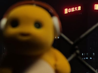 夜晚场景中，前景是一个黄色的毛绒玩偶，戴着红色头带和白色耳罩，面部表情温和，眼睛较大。背景有红色霓虹灯招牌，显示'中国大唐'字样，旁边还有部分'唐'字的霓虹灯光。玩偶位于画面左侧，略微模糊，背景建筑结构隐约可见，整体氛围带有夜晚城市的灯光感。