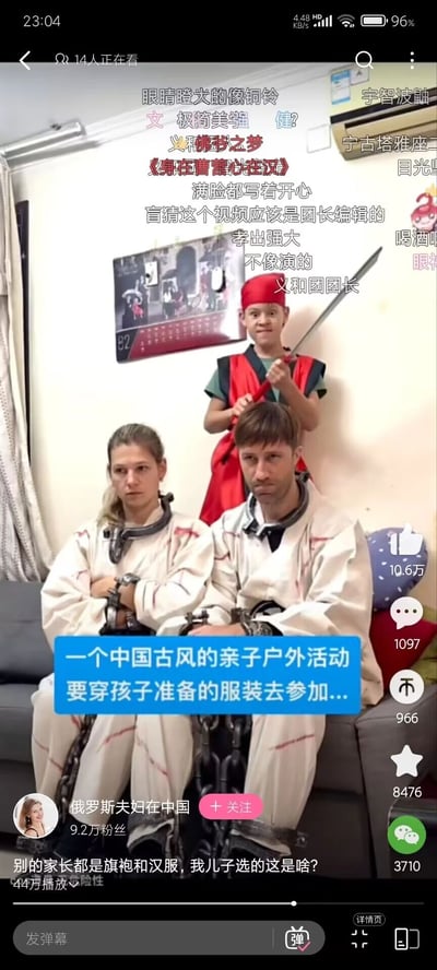 这是一张来自抖音短视频平台的视频截图，由账号“俄罗斯夫妇在中国”发布。画面中，一对在中国生活的俄罗斯夫妇被孩子打扮成古代囚犯的模样：他们穿着带有血痕的白色囚服，颈部和手部戴着枷锁，坐在沙发上，表情无奈又不情愿；他们的孩子则穿着红色的类似刽子手的服装，头戴红帽，手持长刀站在父母身后，表情搞怪。视频字幕显示，他们要参加中国古风亲子户外活动，别的家长都穿旗袍和汉服，孩子却给父母选了这样的服装，形成强烈反差。画面上方布满了网友的玩梗弹幕，比如“义和团团长”“宁古塔雅座”“身在曹营心在汉”等，结合服装造型制造出搞笑的氛围，该视频播放量达44万，互动数据较高，充满了家庭日常的趣味和中外文化碰撞的笑点。