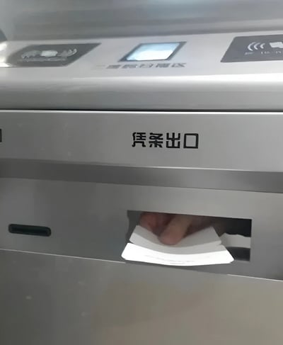 图片近距离拍摄了一台银色自助设备的局部，设备正面中央位置印有黑色文字“凭条出口”，其下方设有一个长方形的出纸口。一只手正从出纸口中取出一张白色的凭条，凭条边缘略微卷曲。设备上部可见一个小型显示屏和几个功能图标，整体呈现出日常自助服务场景的真实感。