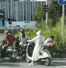 这是一张在城市道路路口拍摄的真实照片，画面中共有四辆电动车停在路口等待通行。最右侧的白色电动车上，骑手身着白色连帽长款外套，后座的孩子身体完全趴在电动车尾部，姿势特殊，未采取安全的乘坐方式，存在交通安全隐患。其余几辆电动车上也有不同的骑行人员，背景可见城市高层建筑、交通信号灯与路边绿植，整体呈现出日常的城市通勤场景，而孩子的特殊姿势成为画面的视觉焦点，引发对电动车出行安全的关注。