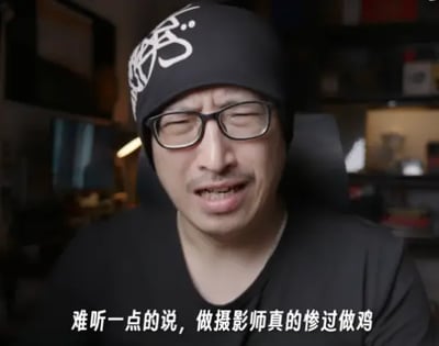 整张图为短视频或直播的截取画面，主体是一名佩戴黑框眼镜、头戴印有白色涂鸦图案黑色冷帽的男性，他眉头紧皱、双眼眯起，面露苦涩无奈的神情，似乎正在吐槽相关内容，画面底部配有白色字幕“难听一点的说，做摄影师真的惨过做鸡”，用极度夸张的表述抒发对摄影师行业辛苦、回报低的不满。