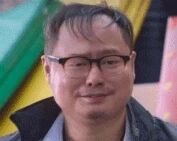 图片中是一位戴着眼镜的男性，短发，面部表情平和，处于静止人像状态。背景包含绿色和粉色等彩色元素，整体氛围偏向日常记录风格。