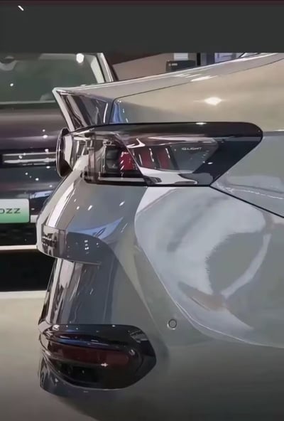 图片为银色汽车尾部的特写镜头，聚焦于现代感十足的尾灯设计，灯组呈现红黑渐变效果，车身表面光滑反光，映出展厅灯光。背景中可见另一辆汽车的部分前脸及车牌（含“DZZ”字样），整体场景推测为汽车展厅或新车发布会现场，突出车辆的精致外观与工业设计美学。