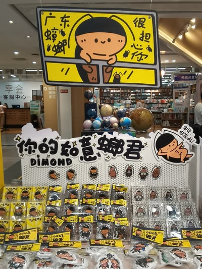 图片展示了一个商店内的蟑螂主题周边商品陈列场景。上方悬挂着黄色背景的卡通招牌，左侧写有“广东蟑螂”及蟑螂简笔画，中间是一个双手托腮的可爱卡通人物形象，右侧配有“很担心你”文字。下方货架上整齐摆放着印有“你的如意螂君 DIAMOND”字样的周边商品，包括多个包装好的徽章和贴纸，商品上印有不同造型的卡通蟑螂图案。背景可见书店或文创店内的书架、地球仪等陈设，整体氛围轻松有趣。