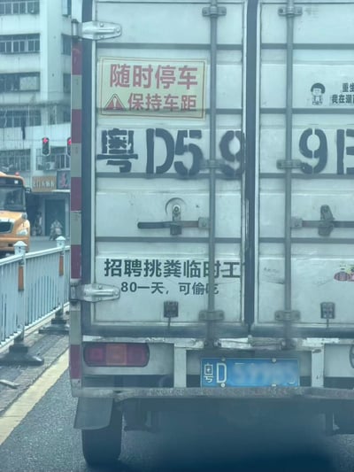 这是一张在城市道路上拍摄的白色厢式货车车尾的实拍照片，车尾上部贴有黄底红边的警示贴纸，印有红色字样“随时停车 保持车距”及警示三角标识，贴纸下方有黑色喷涂的部分车牌信息，开头为粤D59；车尾中部印有黑色的搞笑招聘文字：“招聘挑粪临时工 80一天，可偷吃”，内容荒诞滑稽；车尾底部悬挂粤D开头的蓝色机动车号牌。背景可见城市道路的白色护栏、亮红灯的交通信号灯、路旁的居民楼建筑，以及对面车道行驶的黄色车辆，整体是日常街景中偶然拍到的趣味内容，因招聘启事中离谱的“可偷吃”说明形成笑点。