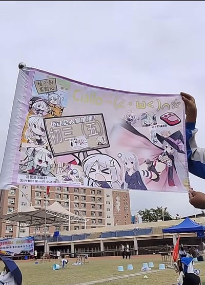 图片展示了在校园运动会现场，有人手持一面设计精美的动漫风格应援旗。旗面主体为白色，印有多个原创动漫少女角色，角色们表情多样，有开心、惊讶、害羞等，动作包括举牌、欢呼、比心等。旗面中央有醒目的文字“运动会大家加油！初三（五）”，周围还有“柚子厨蒸鹅心”“Ciao~(✧ω✧)ﾉ☆”“柚子们赛高！”“这我没法解释因为我只是一只小丛雨”等趣味文字。背景可见学校教学楼、操场及蓝色帐篷，整体氛围充满青春活力与班级凝聚力。