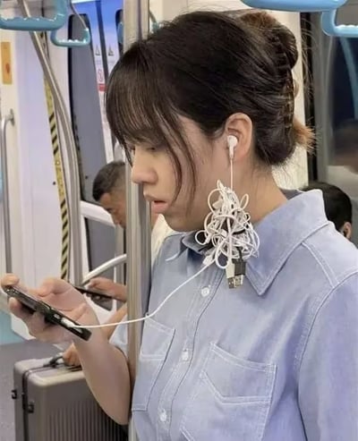 地铁车厢内，一位扎着丸子头、身穿蓝色衬衫的女生正低头专注看手机。她戴着白色耳机，但耳机线在耳后缠成一团乱麻，像个“线团装饰”般垂在脖颈处，完美演绎了“懒得整理耳机线”的摆烂日常。背景中可见其他乘客和地铁扶手，场景真实接地气，让人会心一笑的同时也感同身受。