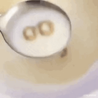 这是一段趣味实拍动图，画面里盛着牛奶的勺子中有两个圈状麦片，外观类似数字“00”，当勺子被提起时，其中一个麦片从勺子边缘掉落到下方的牛奶中，原本的“00”就变成了单个“0”，利用日常早餐食物制造出有趣的视觉小效果，充满生活化的轻松趣味。